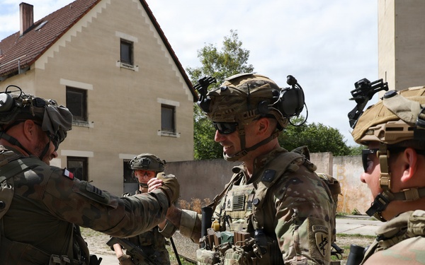 The image shows a group of military personnel in full combat gear interacting in what appears to be a training or operational environment near European-style buildings. Soldiers wearing different national insignia, including what appears to be a French flag patch and U.S. Army Airborne patches, are engaged in a briefing or coordination exchange. The scene suggests a multinational military exercise or joint operation.