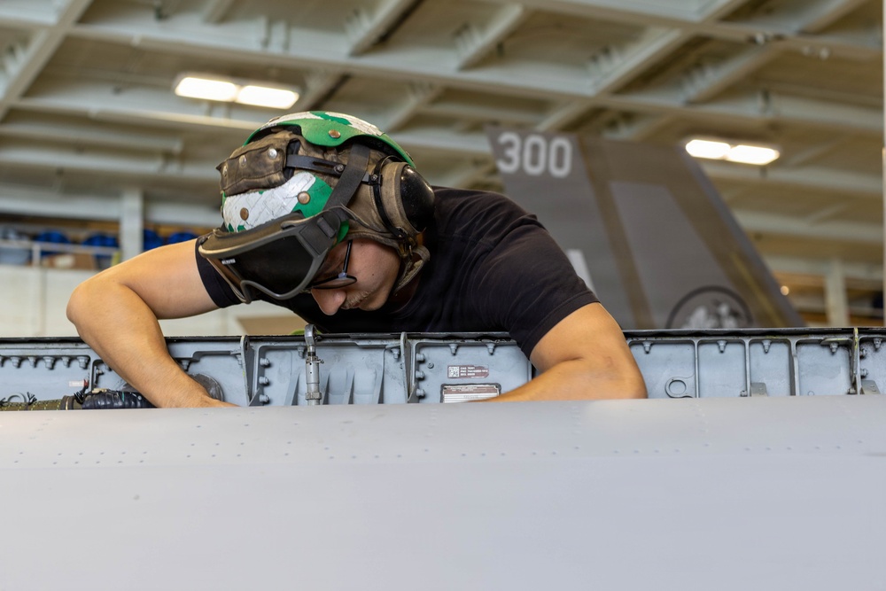 A military aviation technician wearing a green flight deck helmet with hearing protection is performing maintenance or inspection work on an aircraft component, likely a wing or fuselage panel. The work is taking place inside what appears to be a naval vessel's hangar bay, with another aircraft visible in the background bearing the number '300'. The setting and equipment are consistent with U.S. Navy carrier-based aircraft maintenance operations.