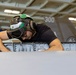 A military aviation technician wearing a green flight deck helmet with hearing protection is performing maintenance or inspection work on an aircraft component, likely a wing or fuselage panel. The work is taking place inside what appears to be a naval vessel's hangar bay, with another aircraft visible in the background bearing the number '300'. The setting and equipment are consistent with U.S. Navy carrier-based aircraft maintenance operations.