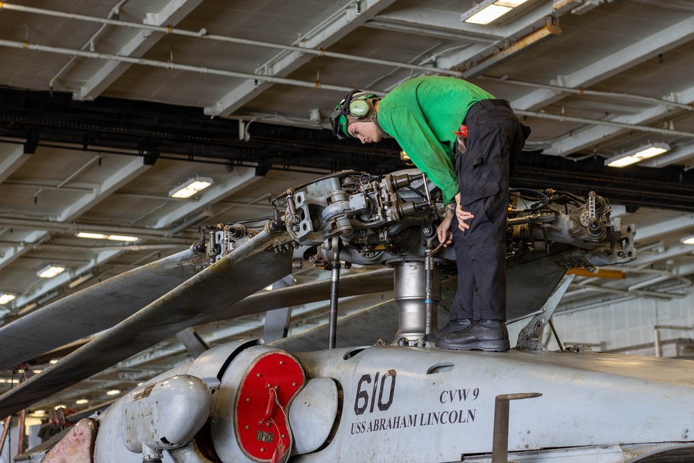 A military aviation technician in a green shirt and black pants is performing maintenance on the rotor head of a helicopter aboard the USS Abraham Lincoln (CVN-72). The aircraft is marked with the number '610' and 'CVW 9' (Carrier Air Wing Nine), indicating it belongs to that carrier air wing. The work is being conducted in the ship's enclosed hangar bay.