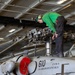 A military aviation technician in a green shirt and black pants is performing maintenance on the rotor head of a helicopter aboard the USS Abraham Lincoln (CVN-72). The aircraft is marked with the number '610' and 'CVW 9' (Carrier Air Wing Nine), indicating it belongs to that carrier air wing. The work is being conducted in the ship's enclosed hangar bay.