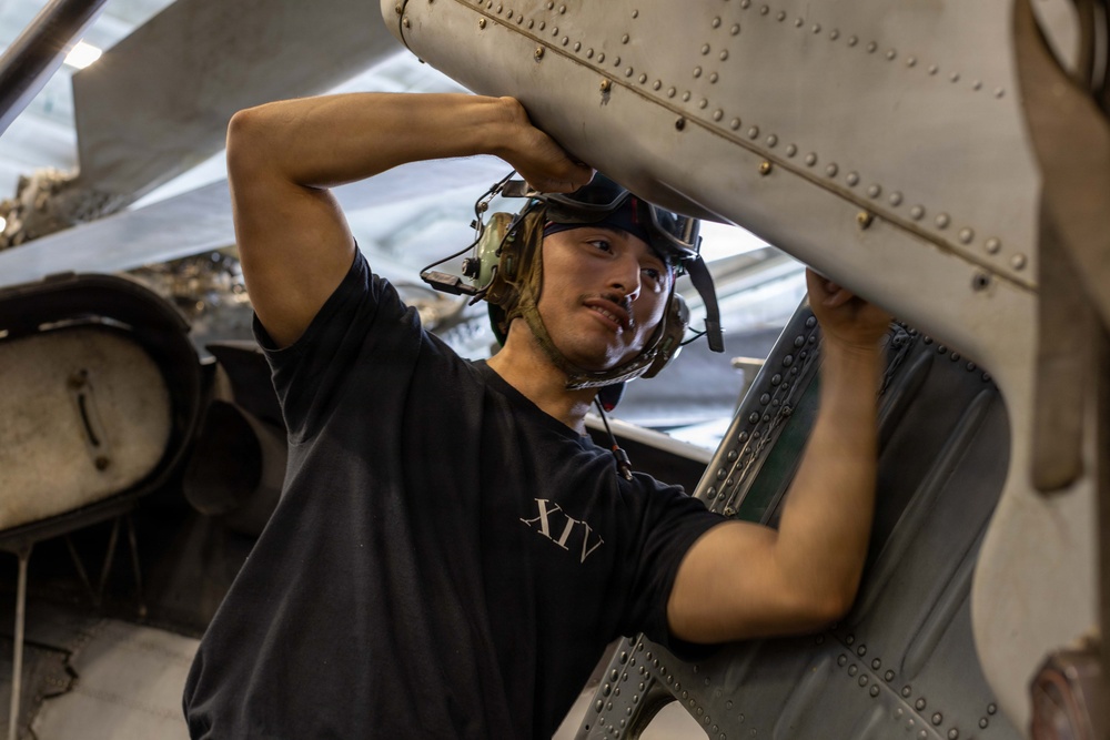 A young male military technician or aviation mechanic is performing maintenance work on an aircraft, likely in a hangar bay aboard a naval vessel. He is wearing a black t-shirt marked with Roman numerals 'XIV' and communication headgear, and is physically manipulating a riveted metal aircraft panel or component. The close-up angle and industrial surroundings suggest an active maintenance operation.