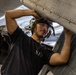 A young male military technician or aviation mechanic is performing maintenance work on an aircraft, likely in a hangar bay aboard a naval vessel. He is wearing a black t-shirt marked with Roman numerals 'XIV' and communication headgear, and is physically manipulating a riveted metal aircraft panel or component. The close-up angle and industrial surroundings suggest an active maintenance operation.