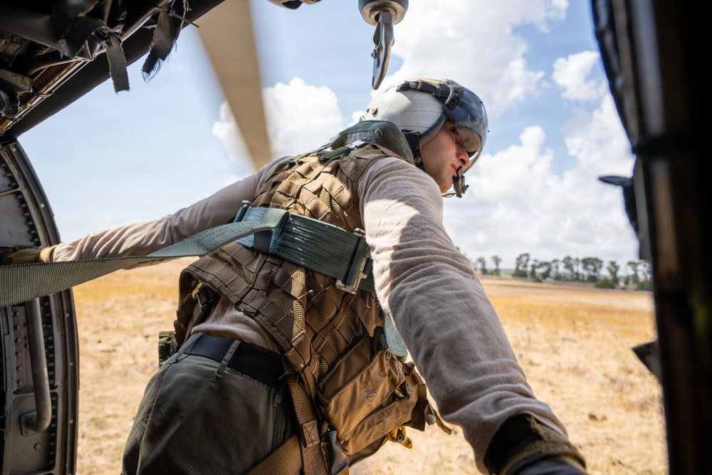 A military or aviation crew member is leaning out of an open helicopter door, scanning the ground below. The individual is wearing a flight helmet with visor, tactical vest, and is secured by a safety harness/strap attached to the aircraft. The background shows an arid, open landscape with sparse trees under a partly cloudy sky.