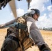 A military or aviation crew member is leaning out of an open helicopter door, scanning the ground below. The individual is wearing a flight helmet with visor, tactical vest, and is secured by a safety harness/strap attached to the aircraft. The background shows an arid, open landscape with sparse trees under a partly cloudy sky.