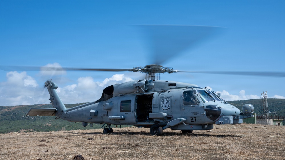 A U.S. Navy military helicopter, marked with the number '27' and 'NAVY' designation, is on the ground with its rotors spinning on an open field. The aircraft appears to be either preparing for takeoff or has just landed at an improvised landing zone. The setting includes hilly terrain, dry grass, and what appears to be a communications or radar installation in the background.
