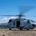 A U.S. Navy military helicopter, marked with the number '27' and 'NAVY' designation, is on the ground with its rotors spinning on an open field. The aircraft appears to be either preparing for takeoff or has just landed at an improvised landing zone. The setting includes hilly terrain, dry grass, and what appears to be a communications or radar installation in the background.