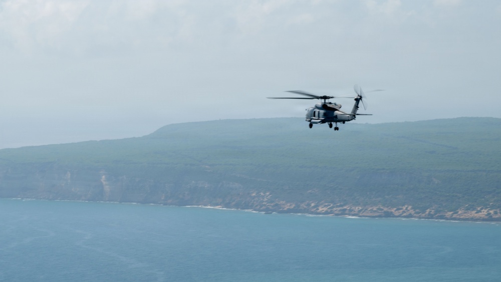 A military helicopter is captured in flight over a coastal area, with a rocky cliff shoreline and blue-green ocean water visible in the background. The helicopter appears to be a naval variant, likely a Sikorsky SH-60 Seahawk or similar platform, painted in a gray military color scheme. The image was taken from an elevated or aerial vantage point with hazy atmospheric conditions.