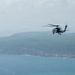 A military helicopter is captured in flight over a coastal area, with a rocky cliff shoreline and blue-green ocean water visible in the background. The helicopter appears to be a naval variant, likely a Sikorsky SH-60 Seahawk or similar platform, painted in a gray military color scheme. The image was taken from an elevated or aerial vantage point with hazy atmospheric conditions.