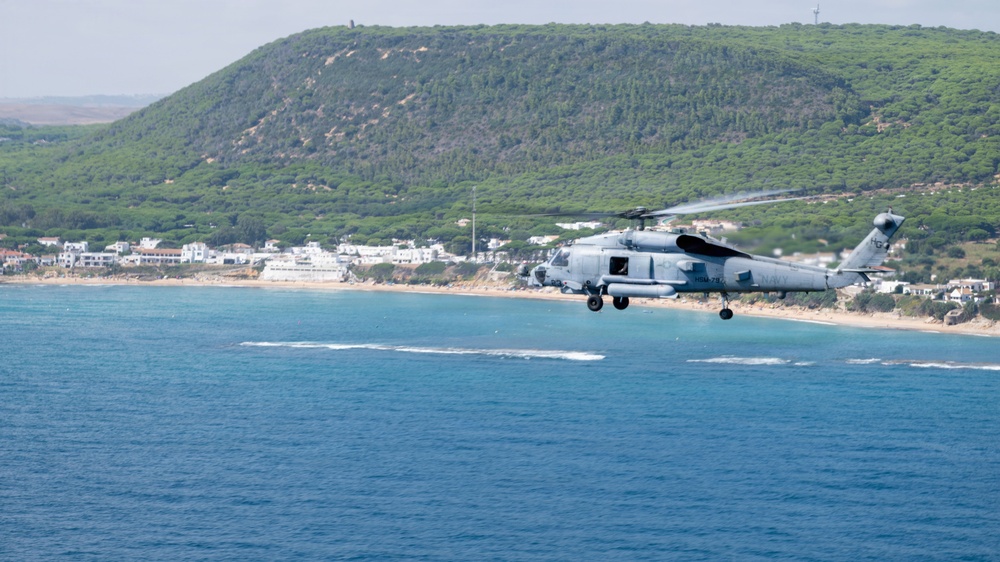 A military helicopter, appearing to be a Sikorsky SH-60 Seahawk or similar naval variant, flies low over a blue coastal body of water near a sandy beach. The helicopter is painted in a light gray naval color scheme and is flying parallel to a shoreline backed by white buildings and a green, tree-covered hill. The rotor wash is visibly disturbing the water surface below the aircraft.