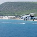 A military helicopter, appearing to be a Sikorsky SH-60 Seahawk or similar naval variant, flies low over a blue coastal body of water near a sandy beach. The helicopter is painted in a light gray naval color scheme and is flying parallel to a shoreline backed by white buildings and a green, tree-covered hill. The rotor wash is visibly disturbing the water surface below the aircraft.