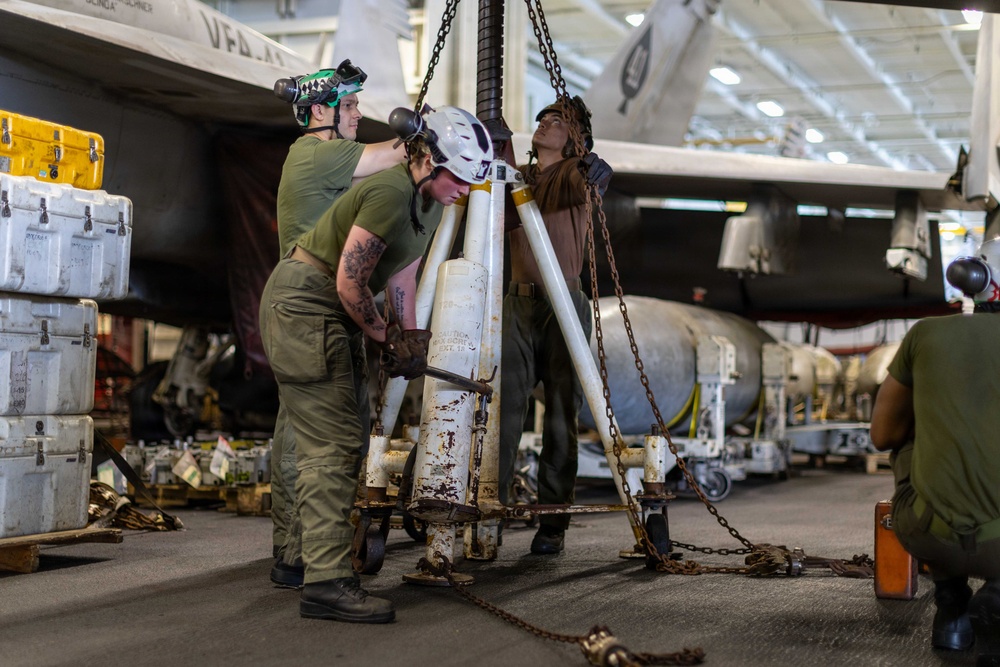 Military personnel in olive drab uniforms are working in what appears to be an aircraft carrier hangar bay, handling ordnance or heavy equipment using a tripod hoist and chain rigging system. Multiple service members wearing helmets and protective gear are collaborating on the lift operation surrounded by storage containers and additional munitions on carts. The setting is a large enclosed hangar with fluorescent lighting and military aircraft visible in the background.