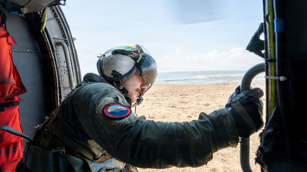 A military or coast guard aircrew member is leaning out of a helicopter door, scanning a sandy beach and ocean coastline. The individual is wearing a flight helmet with a reflective visor and an olive-green flight suit with a unit patch on the sleeve. A red survival vest is visible hanging inside the aircraft cabin to the left.