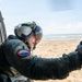 A military or coast guard aircrew member is leaning out of a helicopter door, scanning a sandy beach and ocean coastline. The individual is wearing a flight helmet with a reflective visor and an olive-green flight suit with a unit patch on the sleeve. A red survival vest is visible hanging inside the aircraft cabin to the left.