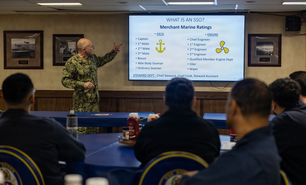 A military officer in U.S. Navy camouflage uniform is presenting a briefing on Merchant Marine Ratings to a group of seated attendees. The presentation slide on the screen explains 'What is an SSO?' and lists Merchant Marine ratings across Deck, Engine, and Steward departments. The setting appears to be a formal conference or briefing room with naval insignia on the chairs.