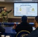 A military officer in U.S. Navy camouflage uniform is presenting a briefing on Merchant Marine Ratings to a group of seated attendees. The presentation slide on the screen explains 'What is an SSO?' and lists Merchant Marine ratings across Deck, Engine, and Steward departments. The setting appears to be a formal conference or briefing room with naval insignia on the chairs.