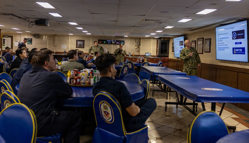 A U.S. Navy briefing or training session is taking place in what appears to be a ship's mess hall or dining facility. Two uniformed Navy personnel are presenting to a seated audience using a projected slideshow displaying 'Navy COOL' (Credentialing Opportunities On-Line) program information. The setting features distinctive blue chairs with Navy insignia emblems.