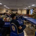 A U.S. Navy briefing or training session is taking place in what appears to be a ship's mess hall or dining facility. Two uniformed Navy personnel are presenting to a seated audience using a projected slideshow displaying 'Navy COOL' (Credentialing Opportunities On-Line) program information. The setting features distinctive blue chairs with Navy insignia emblems.
