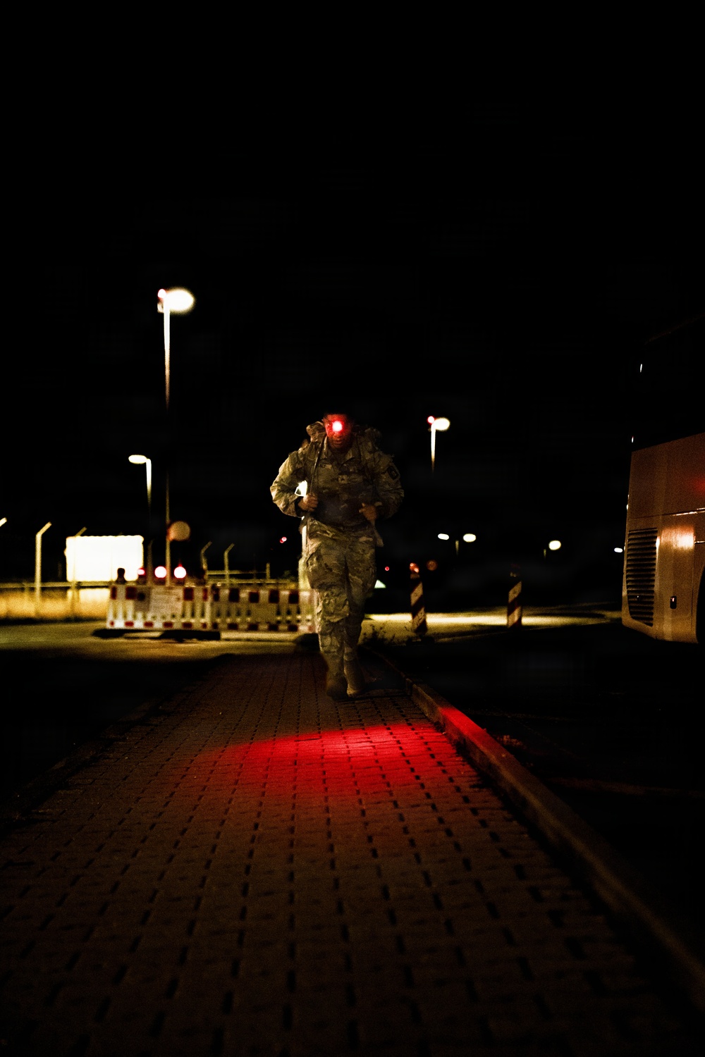 A military service member in camouflage uniform is running or marching at night on a brick-paved surface, carrying a backpack and illuminated by a red light mounted on their head or helmet. The scene appears to be a military installation or checkpoint area, identifiable by road barriers, street lights, and what appears to be a large vehicle on the right. The red light casts a vivid glow on the pavement directly beneath the subject.
