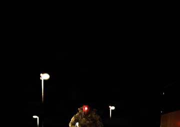 A military service member in camouflage uniform is running or marching at night on a brick-paved surface, carrying a backpack and illuminated by a red light mounted on their head or helmet. The scene appears to be a military installation or checkpoint area, identifiable by road barriers, street lights, and what appears to be a large vehicle on the right. The red light casts a vivid glow on the pavement directly beneath the subject.