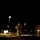 12th Combat Aviation Brigade hosts Danish Flyver March A military service member in camouflage uniform is running or marching at night on a brick-paved surface, carrying a backpack and illuminated by a red light mounted on their head or helmet. The scene appears to be a military installation or checkpoint area, identifiable by road barriers, street lights, and what appears to be a large vehicle on the right. The red light casts a vivid glow on the pavement directly beneath the subject.