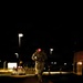 A military service member in camouflage uniform is running or marching at night on a brick-paved surface, carrying a backpack and illuminated by a red light mounted on their head or helmet. The scene appears to be a military installation or checkpoint area, identifiable by road barriers, street lights, and what appears to be a large vehicle on the right. The red light casts a vivid glow on the pavement directly beneath the subject.