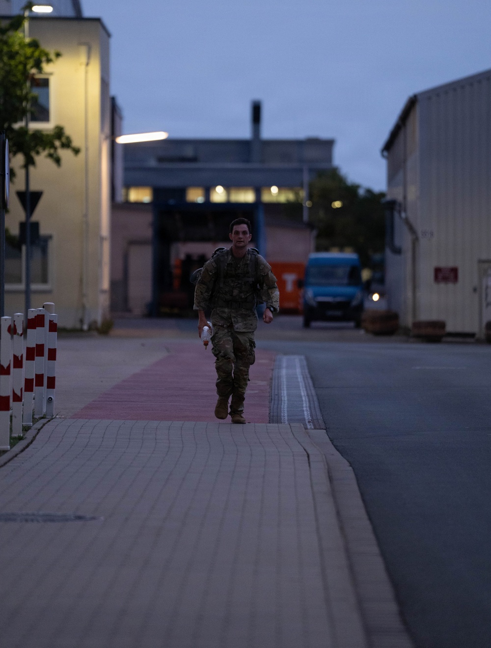 A soldier in camouflage uniform and tactical gear is running or marching at a brisk pace along a paved pathway between buildings during dusk or early dawn. The individual is carrying a backpack and holding what appears to be a water bottle. The setting appears to be a military installation or industrial compound with street lighting illuminating the scene.