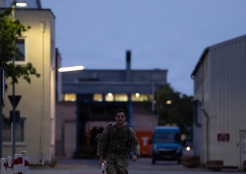 A soldier in camouflage uniform and tactical gear is running or marching at a brisk pace along a paved pathway between buildings during dusk or early dawn. The individual is carrying a backpack and holding what appears to be a water bottle. The setting appears to be a military installation or industrial compound with street lighting illuminating the scene.