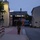 12th Combat Aviation Brigade hosts Danish Flyver March A soldier in camouflage uniform and tactical gear is running or marching at a brisk pace along a paved pathway between buildings during dusk or early dawn. The individual is carrying a backpack and holding what appears to be a water bottle. The setting appears to be a military installation or industrial compound with street lighting illuminating the scene.