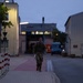 A soldier in camouflage uniform and tactical gear is running or marching at a brisk pace along a paved pathway between buildings during dusk or early dawn. The individual is carrying a backpack and holding what appears to be a water bottle. The setting appears to be a military installation or industrial compound with street lighting illuminating the scene.