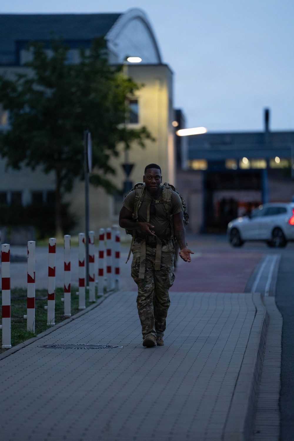 A soldier in camouflage uniform and tactical backpack is walking along a paved pathway during dusk or early morning hours. The setting appears to be a military installation or base, identifiable by the institutional architecture and red-and-white bollards lining the walkway. The individual appears to be conducting a ruck march or physical training exercise.