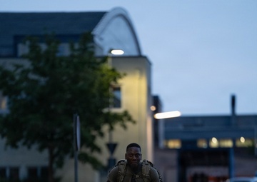 A soldier in camouflage uniform and tactical backpack is walking along a paved pathway during dusk or early morning hours. The setting appears to be a military installation or base, identifiable by the institutional architecture and red-and-white bollards lining the walkway. The individual appears to be conducting a ruck march or physical training exercise.