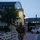 12th Combat Aviation Brigade hosts Danish Flyver March A soldier in camouflage uniform and tactical backpack is walking along a paved pathway during dusk or early morning hours. The setting appears to be a military installation or base, identifiable by the institutional architecture and red-and-white bollards lining the walkway. The individual appears to be conducting a ruck march or physical training exercise.