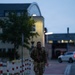 A soldier in camouflage uniform and tactical backpack is walking along a paved pathway during dusk or early morning hours. The setting appears to be a military installation or base, identifiable by the institutional architecture and red-and-white bollards lining the walkway. The individual appears to be conducting a ruck march or physical training exercise.