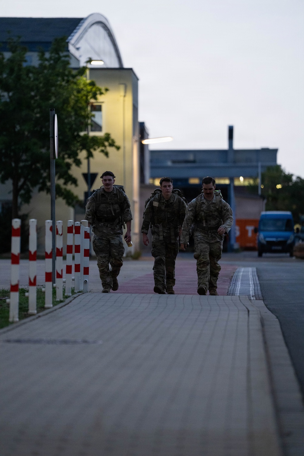 Three military personnel in camouflage uniforms and tactical gear are walking along a paved path on what appears to be a military installation or base. The image is taken during dusk or early morning, with artificial lighting illuminating the background buildings. The soldiers are carrying rucksacks and appear to be conducting a ruck march or returning from a field exercise.