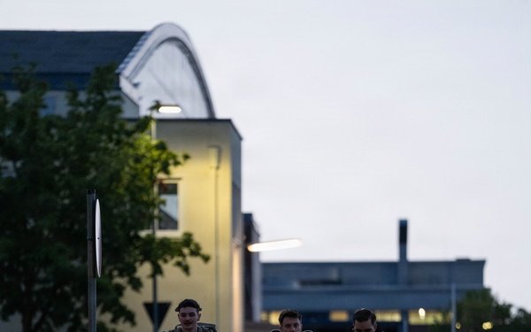 Three military personnel in camouflage uniforms and tactical gear are walking along a paved path on what appears to be a military installation or base. The image is taken during dusk or early morning, with artificial lighting illuminating the background buildings. The soldiers are carrying rucksacks and appear to be conducting a ruck march or returning from a field exercise.