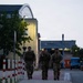 Three military personnel in camouflage uniforms and tactical gear are walking along a paved path on what appears to be a military installation or base. The image is taken during dusk or early morning, with artificial lighting illuminating the background buildings. The soldiers are carrying rucksacks and appear to be conducting a ruck march or returning from a field exercise.