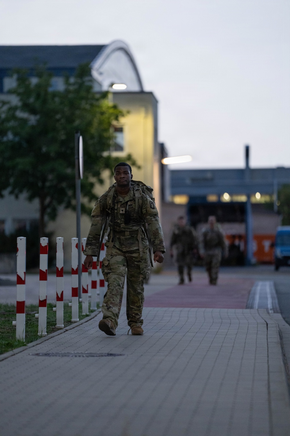 A soldier in full camouflage uniform and tactical gear with a large rucksack walks along a paved pathway during early morning or dusk hours. Additional soldiers are visible in the background following the same route. The setting appears to be a military installation or base, identifiable by the bollards, structured pathways, and institutional buildings.