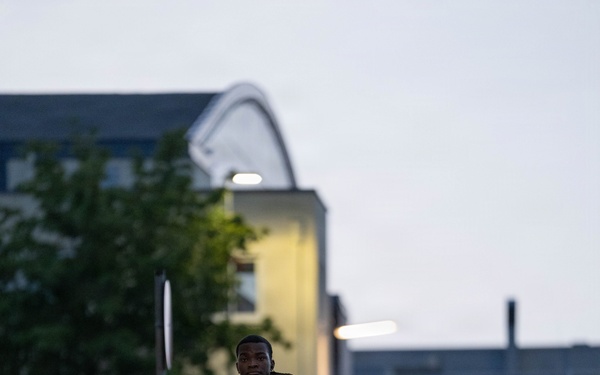 A soldier in full camouflage uniform and tactical gear with a large rucksack walks along a paved pathway during early morning or dusk hours. Additional soldiers are visible in the background following the same route. The setting appears to be a military installation or base, identifiable by the bollards, structured pathways, and institutional buildings.