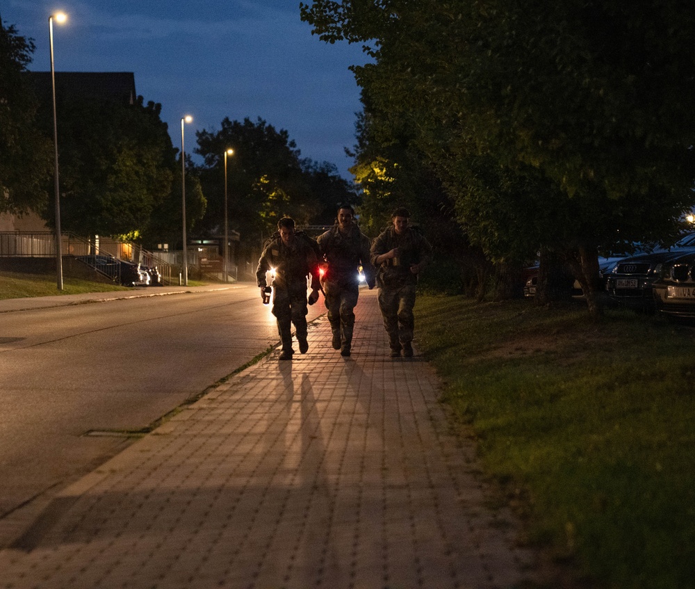 Three individuals in military camouflage uniforms are running together along a paved sidewalk at dusk or nighttime. They appear to be conducting a physical training exercise or ruck march, with one person carrying what appears to be a red light source. The scene is set along a tree-lined road with street lights illuminating the area and parked vehicles visible to the right.