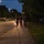 12th Combat Aviation Brigade hosts Danish Flyver March Three individuals in military camouflage uniforms are running together along a paved sidewalk at dusk or nighttime. They appear to be conducting a physical training exercise or ruck march, with one person carrying what appears to be a red light source. The scene is set along a tree-lined road with street lights illuminating the area and parked vehicles visible to the right.