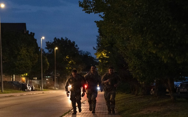 Three individuals in military camouflage uniforms are running together along a paved sidewalk at dusk or nighttime. They appear to be conducting a physical training exercise or ruck march, with one person carrying what appears to be a red light source. The scene is set along a tree-lined road with street lights illuminating the area and parked vehicles visible to the right.
