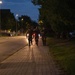 Three individuals in military camouflage uniforms are running together along a paved sidewalk at dusk or nighttime. They appear to be conducting a physical training exercise or ruck march, with one person carrying what appears to be a red light source. The scene is set along a tree-lined road with street lights illuminating the area and parked vehicles visible to the right.