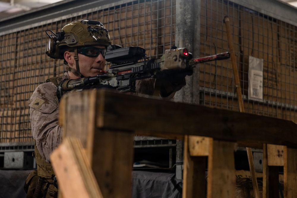 A military service member in full combat gear is aiming a rifle from behind a wooden barricade in what appears to be an indoor training facility. The individual is wearing desert camouflage (MARPAT pattern), a tactical helmet with mounted accessories, and ballistic eyewear. The setting suggests a controlled training environment with wire mesh structures and wooden barriers visible in the background.