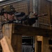 A military service member in full combat gear is aiming a rifle from behind a wooden barricade in what appears to be an indoor training facility. The individual is wearing desert camouflage (MARPAT pattern), a tactical helmet with mounted accessories, and ballistic eyewear. The setting suggests a controlled training environment with wire mesh structures and wooden barriers visible in the background.