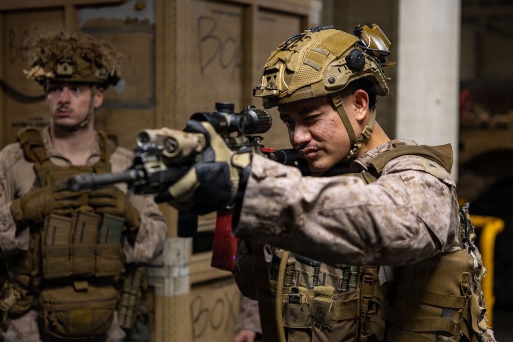 Two military personnel in full combat gear are conducting what appears to be a tactical training exercise or operation in an indoor environment. The foreground soldier is actively aiming a rifle equipped with an optic scope, while the second soldier stands in a ready position behind him. Both wear MARPAT (Marine pattern) camouflage uniforms, suggesting U.S. Marine Corps affiliation.