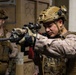 Two military personnel in full combat gear are conducting what appears to be a tactical training exercise or operation in an indoor environment. The foreground soldier is actively aiming a rifle equipped with an optic scope, while the second soldier stands in a ready position behind him. Both wear MARPAT (Marine pattern) camouflage uniforms, suggesting U.S. Marine Corps affiliation.