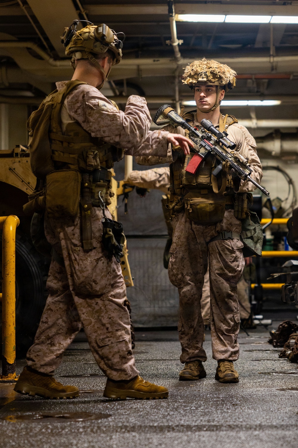 Two military personnel in MARPAT desert camouflage uniforms are shown in what appears to be the well deck or vehicle bay of a naval vessel. One service member appears to be conducting an equipment inspection or pre-mission briefing with the other, who is holding a rifle with a red safety flag attached. Both are in full combat gear including tactical vests, helmets with camouflage netting, and combat boots.