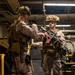 Two military personnel in MARPAT desert camouflage uniforms are shown in what appears to be the well deck or vehicle bay of a naval vessel. One service member appears to be conducting an equipment inspection or pre-mission briefing with the other, who is holding a rifle with a red safety flag attached. Both are in full combat gear including tactical vests, helmets with camouflage netting, and combat boots.