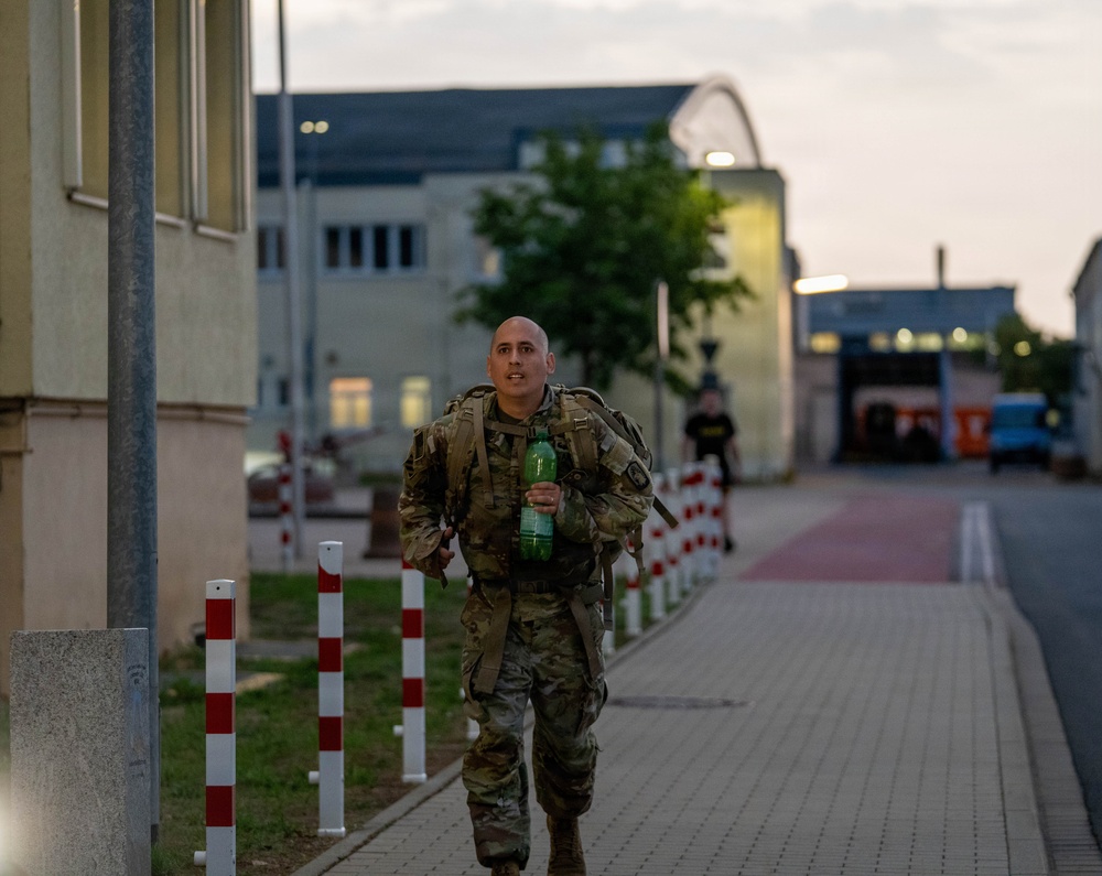 A bald male soldier in MultiCam/OCP camouflage uniform is running or fast-walking along a paved pathway on what appears to be a military installation. He is carrying a tactical backpack/rucksack and holding a green plastic water bottle in his right hand. The setting appears to be dusk or early morning, with institutional buildings and red-and-white bollards lining the path in the background.